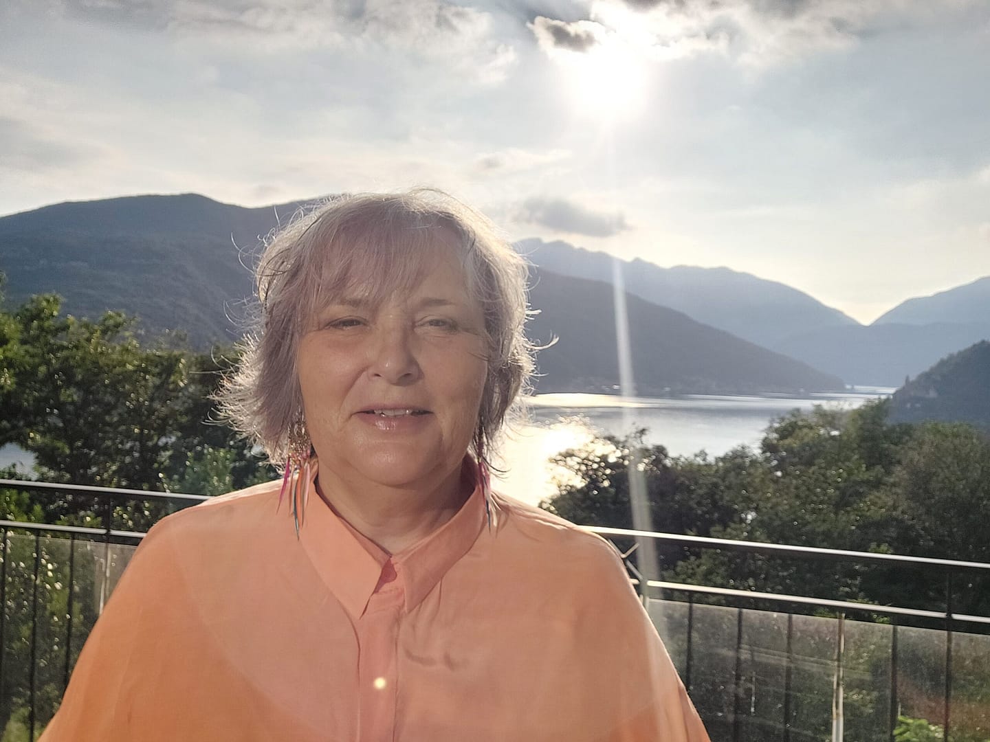 Portrait einer Frau mit silbergrauen Haaren in lachsfarbener Bluse, aufgenommen auf einer sonnigen Terrasse mit Blick auf See und Berge im Hintergrund.