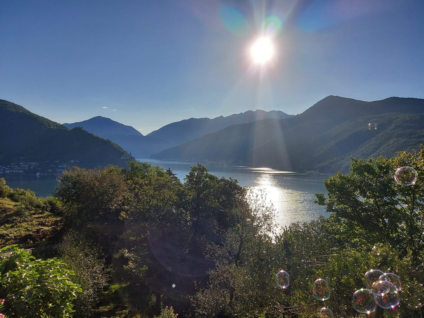 Sonnenaufgang über einem ruhigen See im Gebirge, Lichtreflexe auf dem Wasser, umgeben von grüner Natur und Bergen.