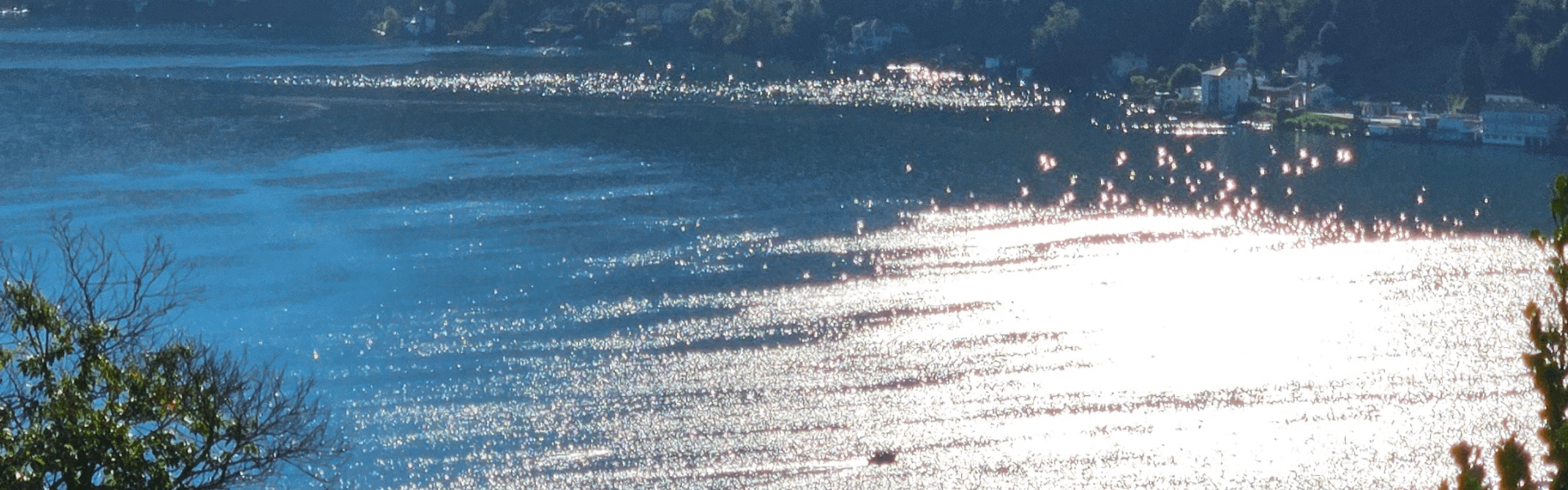 Sonnenlicht glitzert auf türkisblauem Wasser des Lago di Lugano mit Uferpromenade und Bergen im Hintergrund