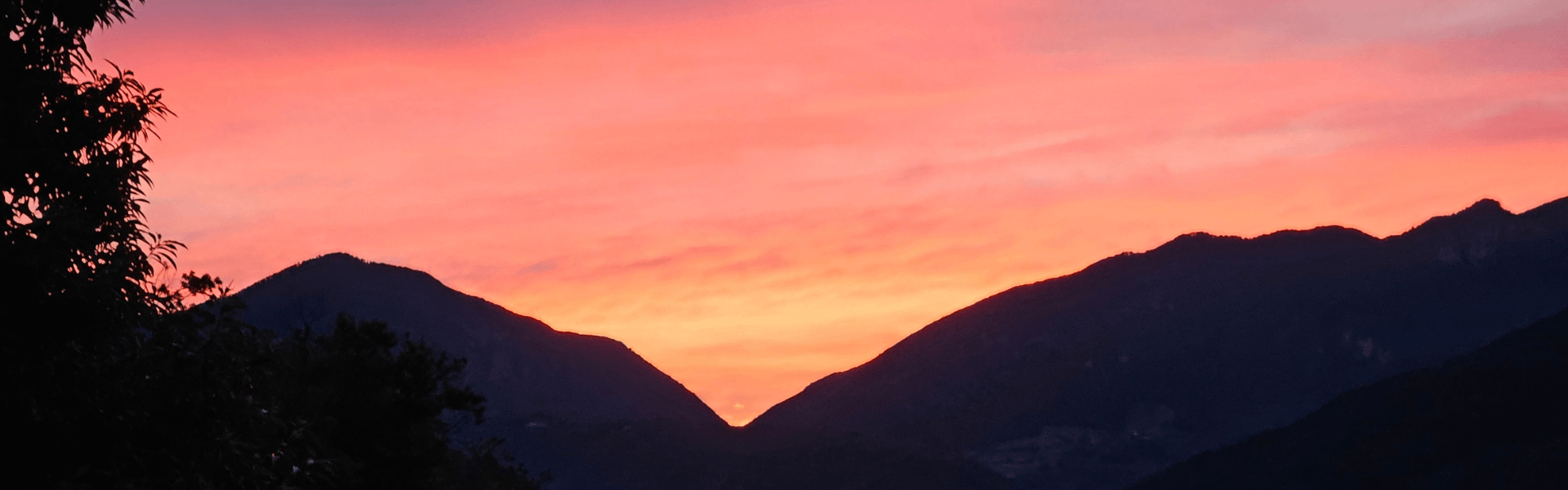 Leuchtender Sonnenuntergang in kräftigem Rosa und Orange zwischen zwei Bergspitzen – intensiver Himmel über ruhiger Landschaft.