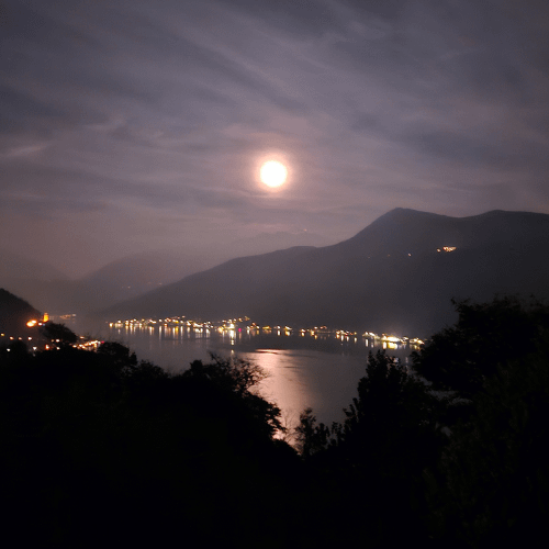 Vollmond über dem Luganersee bei Nacht, Bergsilhouetten im Hintergrund, Stadtlichter spiegeln sich im Wasser, Baumwipfel im Vordergrund